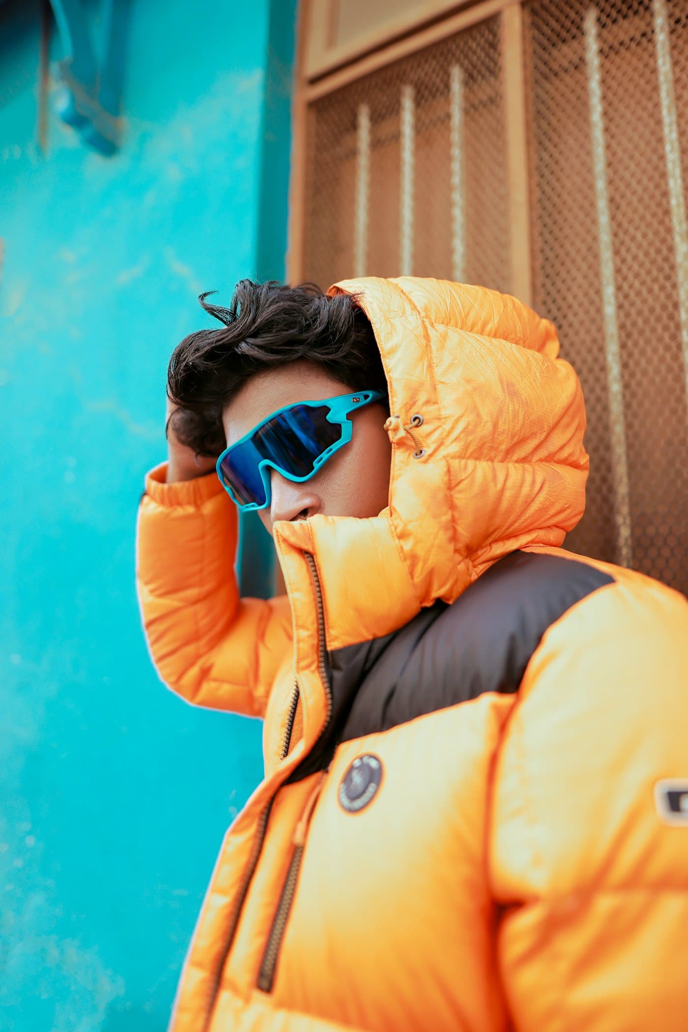 A creator in an orange puffer jacket and blue sunglasses against a turquoise wall